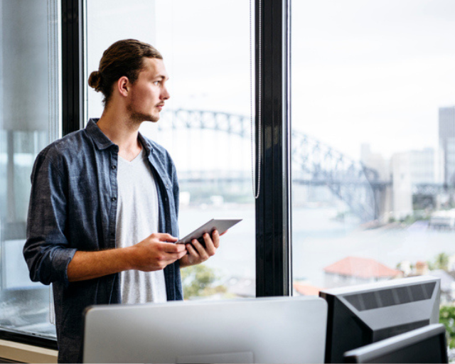 man holding tablet looking outside large window with harbour bridge in background